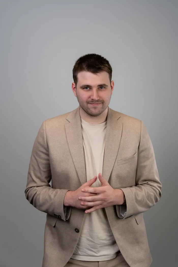 Tomass Bērziņš in a beige blazer and cream T-shirt, hands clasped, against a grey studio background
