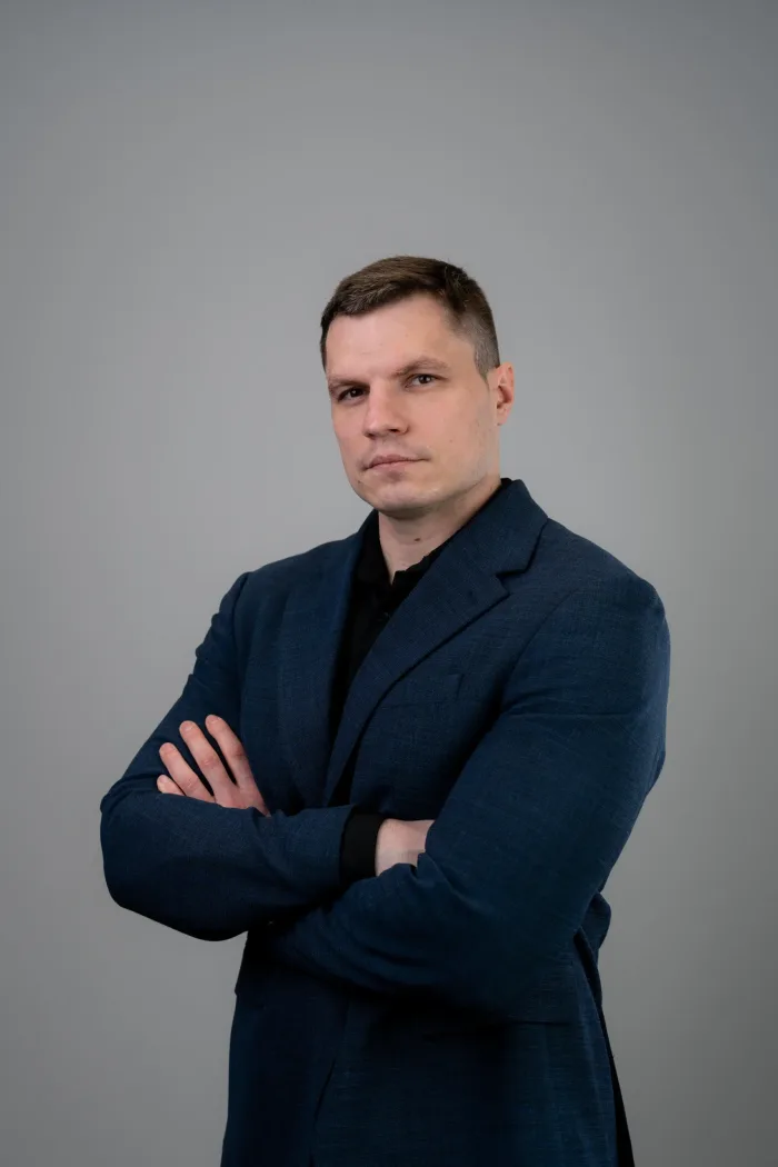 Valdis Strautiņš in a navy blazer and dark shirt, arms crossed, against a grey studio background